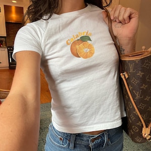 May include: White t-shirt with a graphic of an orange and the word "Calabria" printed on the front. The person is wearing blue jeans and carrying a brown leather handbag.