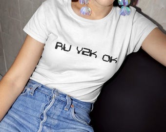 May include: A woman wearing a white t-shirt with the text "RU Y2K OK" printed on it. She is also wearing blue jeans and iridescent earrings.