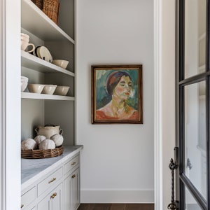 May include: A white kitchen pantry with white shelves, a white countertop, and white cabinets. The shelves are filled with white bowls and a wicker basket filled with white gourds. A framed painting of a woman with a red scarf hangs on the wall.