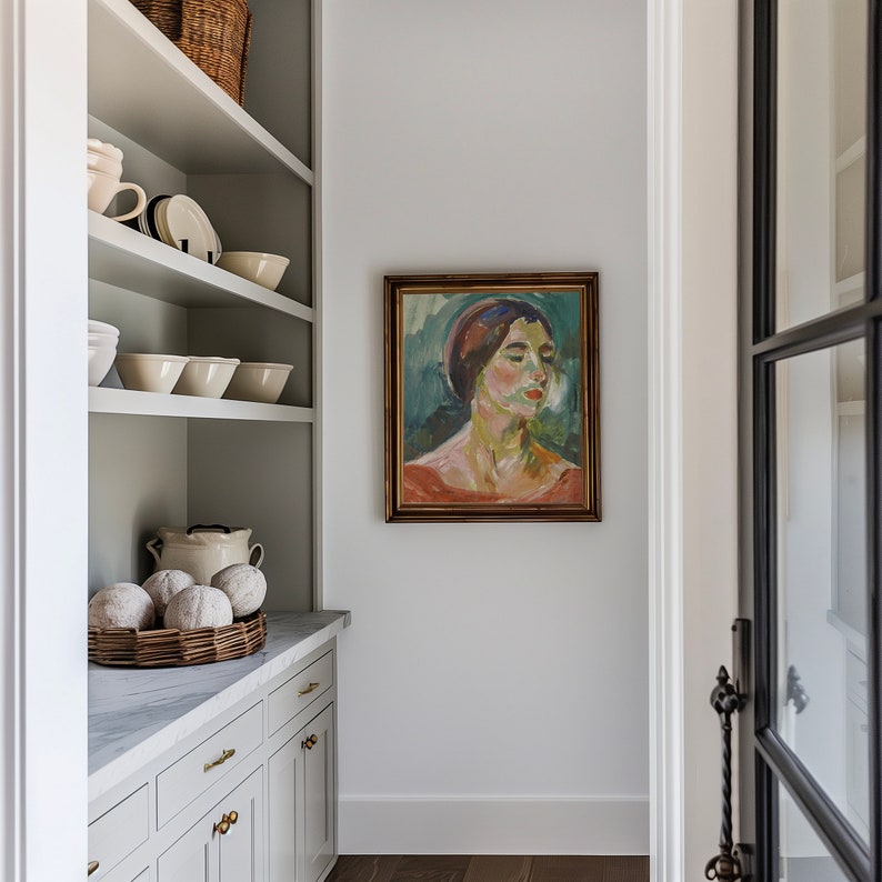 May include: A white kitchen pantry with white shelves, a white countertop, and white cabinets. The shelves are filled with white bowls and a wicker basket filled with white gourds. A framed painting of a woman with a red scarf hangs on the wall.