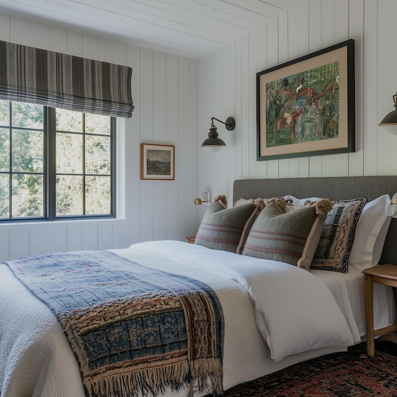 May include: A bedroom with white paneled walls, a window with a striped shade, and a bed with a gray headboard. The bed has a white comforter, a blue and brown patterned throw, and decorative pillows. Artwork hangs on the wall.