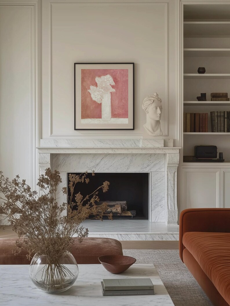 May include: A white marble fireplace with a black fireplace insert. A brown couch is in the foreground with a vase of dried flowers on a white marble coffee table. A framed abstract painting with a pink background hangs above the fireplace.