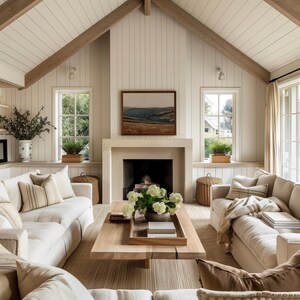 May include: A cozy living room with a neutral color palette. Features include a light wood coffee table, cream-colored sofas, a fireplace with a landscape painting, and large windows. The room is decorated with plants and woven baskets, creating a warm and inviting atmosphere.