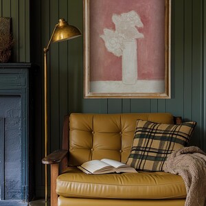 May include: A gold floor lamp with a white shade sits next to a brown leather sofa with a plaid pillow and a book on the seat. A framed print of a white flower arrangement in a vase against a pink background hangs on the wall.