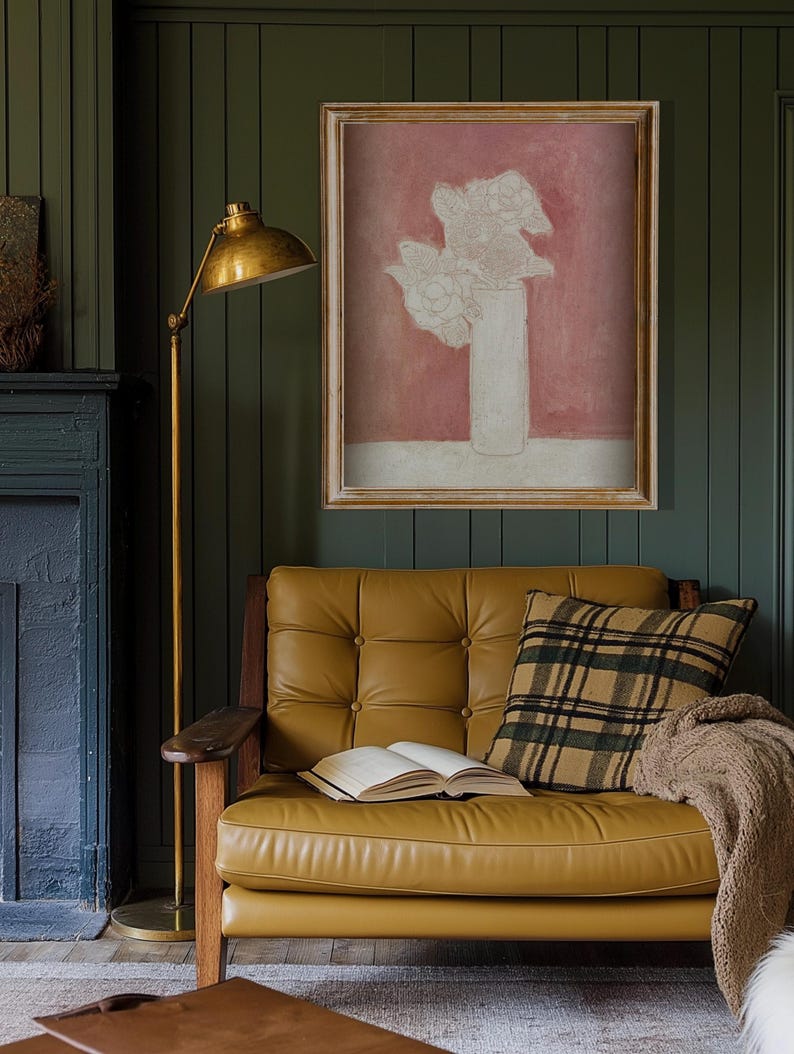 May include: A gold floor lamp with a white shade sits next to a brown leather sofa with a plaid pillow and a book on the seat. A framed print of a white flower arrangement in a vase against a pink background hangs on the wall.
