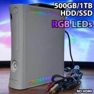 May include: A light grey Xbox 360 console with RGB LED lights and the text "500GB/1TB HDD/SSD RGB LEDs". The console features a power button and a disc tray. Cables with red, white, and yellow connectors are visible.
