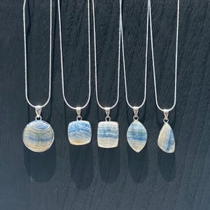 May include: A display of six silver necklaces with blue and white stone pendants. The pendants have various shapes, including round, square, and teardrop. The necklaces are arranged on a black display stand, with a pool in the background.