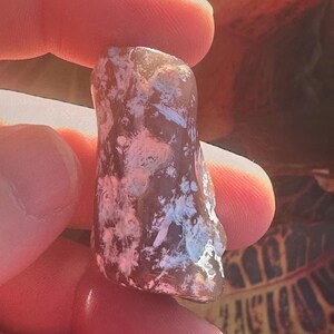 May include: A translucent, oblong gemstone with a mix of brown, pink, and white hues. The stone is held between fingers, showcasing its natural, irregular shape and polished surface. The background features blurred foliage.