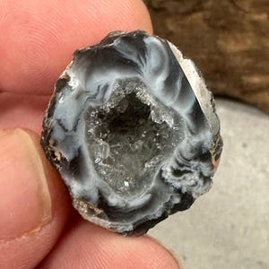 May include: A small, circular agate geode featuring a hollow center filled with sparkling, translucent quartz crystals. The outer rim displays concentric bands of deep black and opaque white, creating a marbled, swirling pattern around the crystal core.