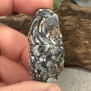 May include: A small, cylindrical stone container with a two-part design. The surface features a complex, natural pattern of dark brown, black, and white swirls with textured, mineral-like inclusions throughout the exterior of the stone.