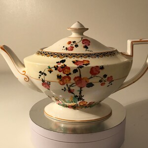 May include: A white teapot with a floral design in shades of pink, orange, and green. The teapot has a round body with a flared spout and a handle that curves up and over the top. The teapot is sitting on a silver pedestal.