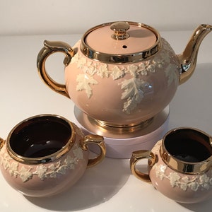 May include: A pink tea set with gold trim and white floral designs. The set includes a teapot, sugar bowl, and creamer.