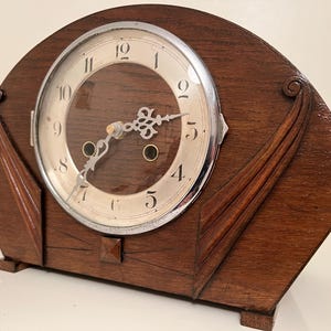 May include: A vintage wooden clock with a silver-toned face and hands. The clock has a dark brown, art deco-style wooden frame with decorative accents. The clock face displays the numbers 1 through 12.