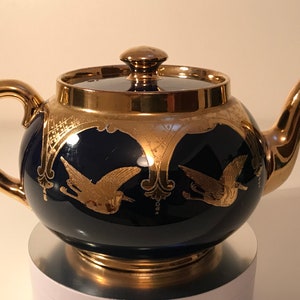 May include: A cobalt blue teapot with gold accents and a design of flying birds. The teapot has a gold lid and a gold handle.
