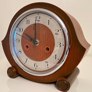May include: A vintage wooden clock with a round face and a brown wooden frame. The clock face has a silver border and black numerals. The clock hands point to 12 o'clock. The clock has two brown cylindrical feet.