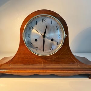May include: A wooden mantel clock with a brass face and black hands. The clock has a Roman numeral clock face and is set on a wooden base.