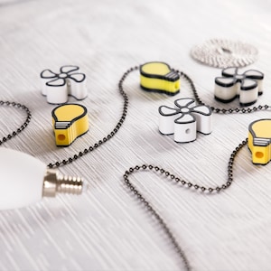 May include: A set of five light switch pull chain extensions. The extensions are made of plastic and feature different designs, including a light bulb, a fan, and a flower. The extensions are attached to black chains.