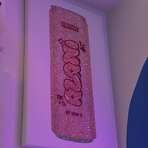 K&ouml;nnte beinhalten: Ein pinkfarbenes, mit Strass besetztes Kunstwerk, das eine Limonadendose darstellt. Das Wort "Frazz" ist in einer Bubble-Schrift geschrieben, mit den Worten "Himade" und "By Kim K" dar&uuml;ber und darunter. Das Kunstwerk ist auf einer wei&szlig;en Leinwand montiert.
