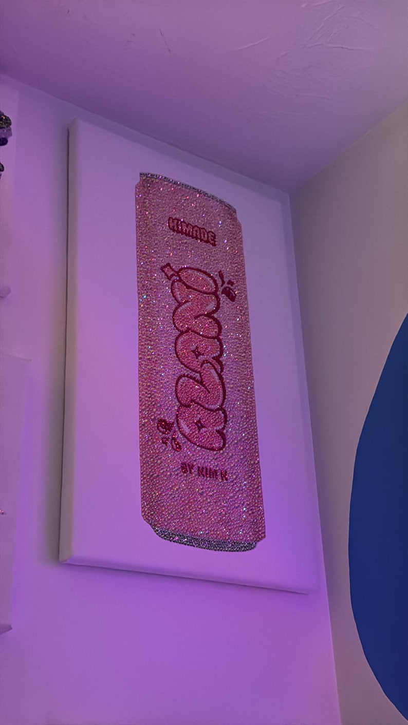 K&ouml;nnte beinhalten: Ein pinkfarbenes, mit Strass besetztes Kunstwerk, das eine Limonadendose darstellt. Das Wort "Frazz" ist in einer Bubble-Schrift geschrieben, mit den Worten "Himade" und "By Kim K" dar&uuml;ber und darunter. Das Kunstwerk ist auf einer wei&szlig;en Leinwand montiert.