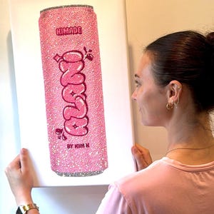 K&ouml;nnte beinhalten: Rosa Leinwandkunst mit einem glitzernden Soda-Dosen-Design. Die Dose tr&auml;gt das Wort "Zano" in einer Blasen-Schriftart, mit dem Text "HIMADE" und "BY KIM K" dar&uuml;ber und darunter. Die Dose ist mit Glitzer bedeckt.