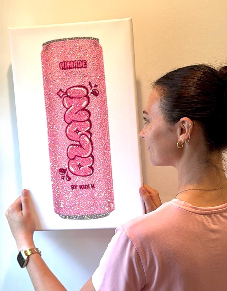 K&ouml;nnte beinhalten: Rosa Leinwandkunst mit einem glitzernden Soda-Dosen-Design. Die Dose tr&auml;gt das Wort "Zano" in einer Blasen-Schriftart, mit dem Text "HIMADE" und "BY KIM K" dar&uuml;ber und darunter. Die Dose ist mit Glitzer bedeckt.
