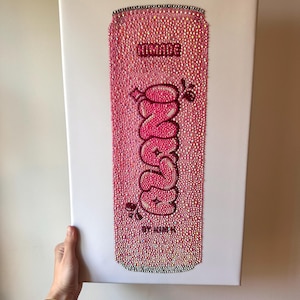 K&ouml;nnte beinhalten: Rosa Kunstwerk einer mit Strasssteinen bedeckten Limonadendose. Das Wort "NINADE" steht oben, mit "ONZE" in einem Bubble-Letter-Stil in der Mitte. Der Text "BY KIM K" steht unten. Das Kunstwerk befindet sich auf einer wei&szlig;en Leinwand.