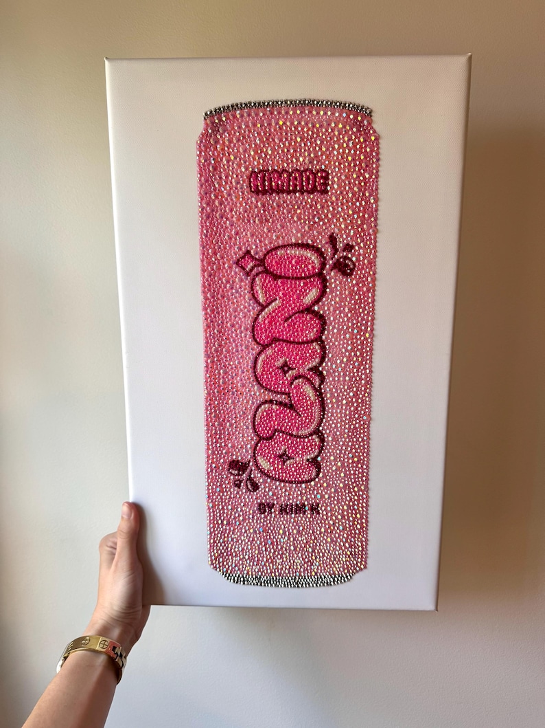 K&ouml;nnte beinhalten: Rosa Kunstwerk einer mit Strasssteinen bedeckten Limonadendose. Das Wort "NINADE" steht oben, mit "ONZE" in einem Bubble-Letter-Stil in der Mitte. Der Text "BY KIM K" steht unten. Das Kunstwerk befindet sich auf einer wei&szlig;en Leinwand.