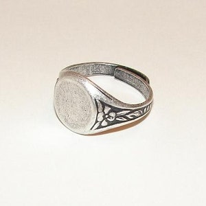 May include: A silver ring with a round, blank face and a floral design on the band.