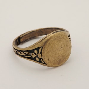 Puede incluir: Un anillo de color dorado con una parte superior ovalada lisa y un diseño floral en la banda.
