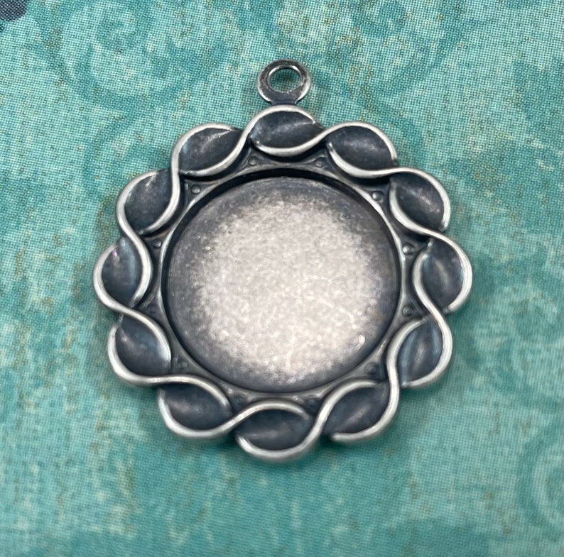 May include: A silver pendant featuring a round, smooth, blank surface encircled by a decorative, twisted, rope-like border. The pendant has a loop at the top for attaching a chain.
