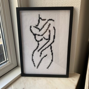May include: A black and white cross-stitch artwork depicting a stylized human figure in a minimalist design. The artwork is framed in a black frame and displayed on a white marble surface.