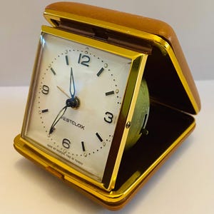 May include: A Westclox travel alarm clock in a tan folding case with gold-tone trim. The square clock face features a white background, black Arabic numerals at the 3, 6, 9, and 12 positions, black hands, and the text Westclox.
