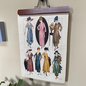 May include: A vintage-style wall hanging featuring a printed illustration of seven figures wearing various dresses and hats from the early 20th century. The print is held by a dark brown wooden frame with a black metal hanger attached to the top.