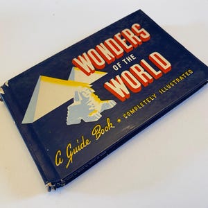 May include: A vintage guide book titled "Wonders of the World" with a navy blue cover. The title is in red and white lettering. The cover features an illustration of a mountain and a plane. The text "A Guide Book * COMPLETELY ILLUSTRATED" is also visible.