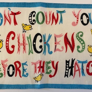May include: A rectangular, off-white linen towel with a blue border. The towel features the phrase "DON'T COUNT YOUR CHICKENS BEFORE THEY HATCH" in colorful, whimsical lettering, along with illustrations of yellow chicks. The artist's signature is in the bottom right corner.