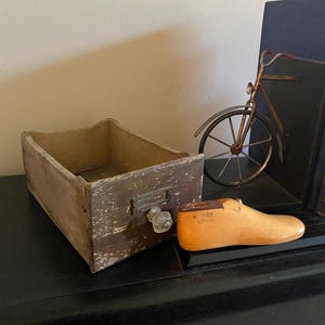 May include: A weathered wooden drawer with a clear glass knob and metal label holder, a light-colored wooden shoe last with the text W 6 5S OCT 1924, and a dark metal bicycle-shaped bookend standing next to a dark blue book.