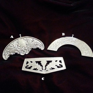 Puede incluir: Tres accesorios de metal plateado etiquetados como A, B y C. El artículo A es una pieza semicircular con grabados florales ornamentados. El artículo B es una pieza semicircular con un patrón de nudo celta. El artículo C es una pieza rectangular con diseños de hojas recortadas.