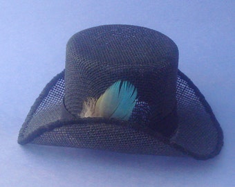 Puede incluir: Un sombrero negro con una copa cilíndrica estructurada y un ala curva, que presenta una textura de malla tejida en toda su superficie. Una pluma decorativa con tonos turquesa brillante y beige suave se inserta en la cinta negra de la base.