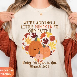 May include: Cream-colored t-shirt with a pumpkin graphic and the text "WE'RE ADDING A LITTLE PUMPKIN TO OUR PATCH!!" The graphic includes three orange pumpkins and fall leaves. The text "Baby Morgan is due March 2025" is also printed on the shirt.
