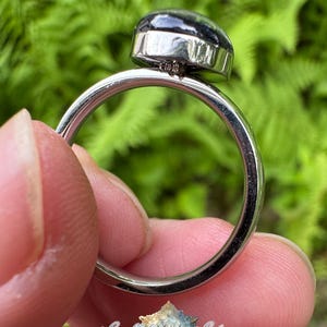 May include: A silver-toned metal ring featuring a smooth, dark, rounded cabochon stone set in a bezel. The ring band is plain and polished. A small logo with the text "Snowflakes & Spiders" and a spider graphic is visible at the bottom.