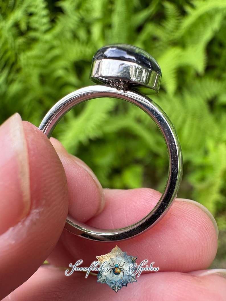 May include: A silver-toned metal ring featuring a smooth, dark, rounded cabochon stone set in a bezel. The ring band is plain and polished. A small logo with the text "Snowflakes & Spiders" and a spider graphic is visible at the bottom.