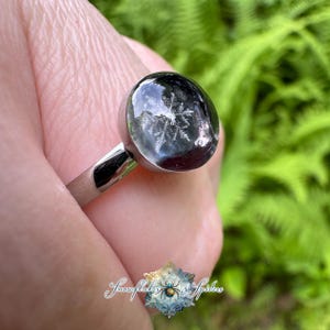 May include: A silver-toned ring featuring a smooth, rounded, dark-colored cabochon setting. Embedded within the translucent, dark dome is a clear, white, six-pointed snowflake pattern. The band is metallic and polished with a simple, clean design.