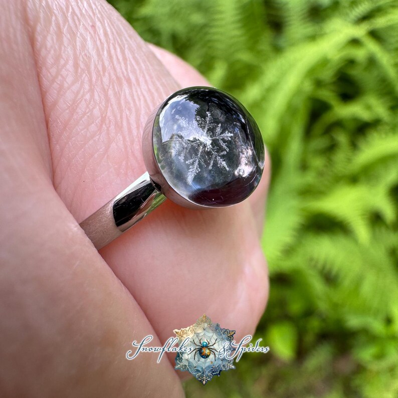 May include: A silver-toned ring featuring a smooth, rounded, dark-colored cabochon setting. Embedded within the translucent, dark dome is a clear, white, six-pointed snowflake pattern. The band is metallic and polished with a simple, clean design.