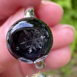 May include: A silver ring featuring a circular, domed black resin cabochon with a white, intricate snowflake design embedded inside. The surface of the resin is smooth and reflective, showcasing the detailed crystalline structure of the snowflake.