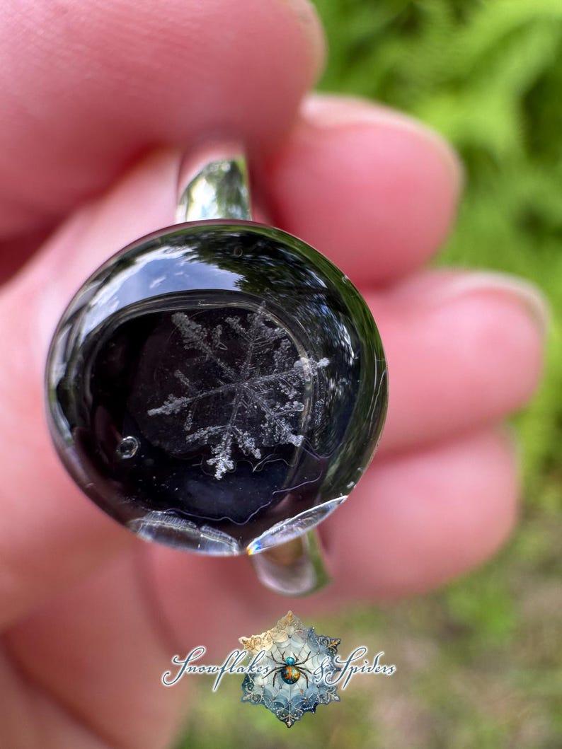 May include: A silver ring featuring a circular, domed black resin cabochon with a white, intricate snowflake design embedded inside. The surface of the resin is smooth and reflective, showcasing the detailed crystalline structure of the snowflake.