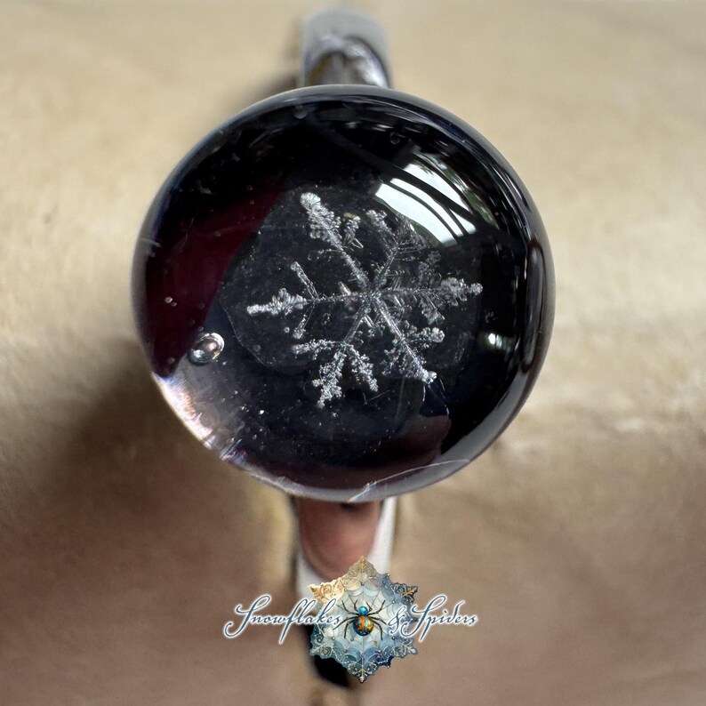 May include: A clear, spherical glass paperweight featuring a detailed, white, six-pointed snowflake design suspended within the center. The object has a smooth, polished surface with a small air bubble visible near the edge of the sphere.