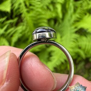 May include: A silver-toned metal ring featuring a smooth, dark, rounded cabochon stone set in a polished bezel. The band is thin and circular, showcasing a clean, metallic finish that reflects light along its curved surface.