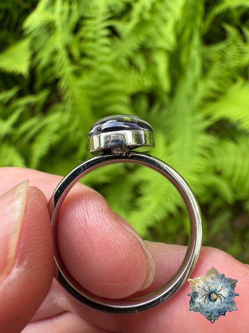 May include: A silver-toned metal ring featuring a smooth, dark, rounded cabochon stone set in a polished bezel. The band is thin and circular, showcasing a clean, metallic finish that reflects light along its curved surface.