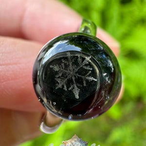 May include: A circular glass ring featuring a dark, translucent dome with a detailed white snowflake pattern embedded inside. The ring has a smooth, polished finish and a clear glass band. Below the ring, a logo reads Snowflakes & Spiders.