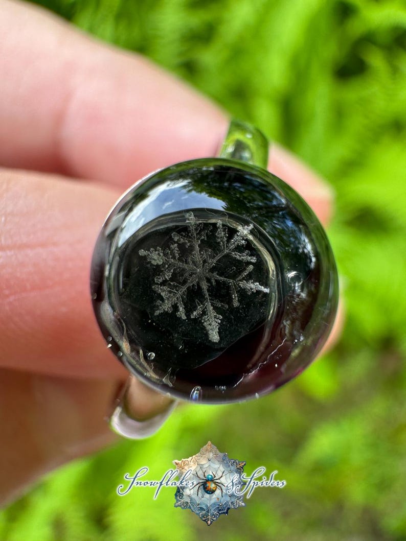 May include: A circular glass ring featuring a dark, translucent dome with a detailed white snowflake pattern embedded inside. The ring has a smooth, polished finish and a clear glass band. Below the ring, a logo reads Snowflakes & Spiders.